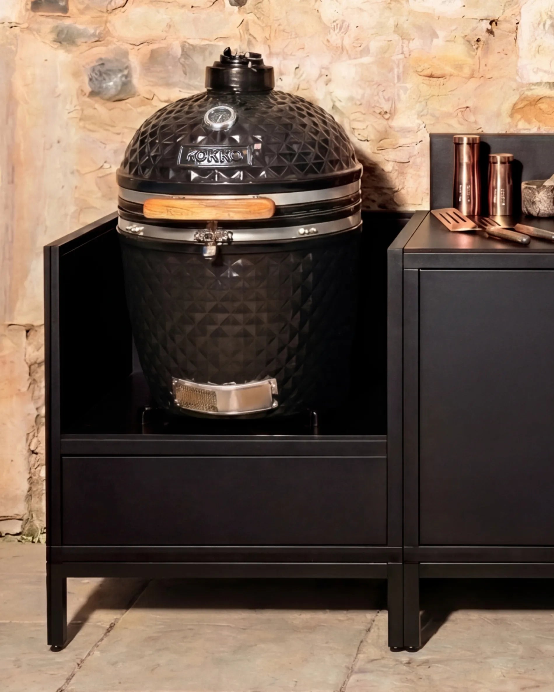 Zwarte Kokko kamado barbecue geplaatst in PILOTIS-ombouw met koperen accessoires, voor buitenkoken