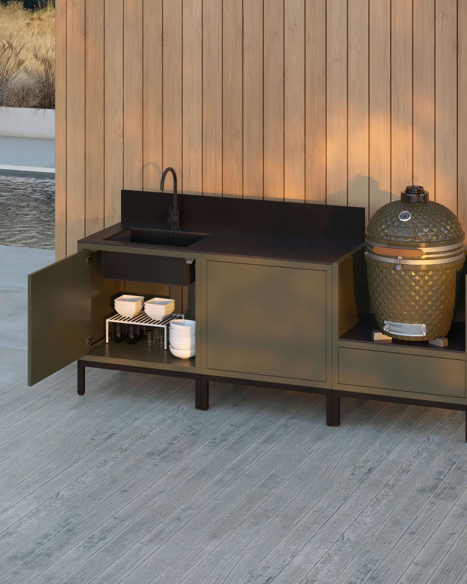Buitenkeuken op palen met 3 modules voor kamado, geïntegreerde spoelbak, meubel in olijfgroen met zwart werkblad en achterwand, linker moduledeur open met zicht op binnenopbergruimte, spoelbak met kraan links, twee gesloten modules in het midden en geïntegreerde kamado in de rechter zijmodule, constructie op zwarte poten, geplaatst buiten tegen een houten gevel.