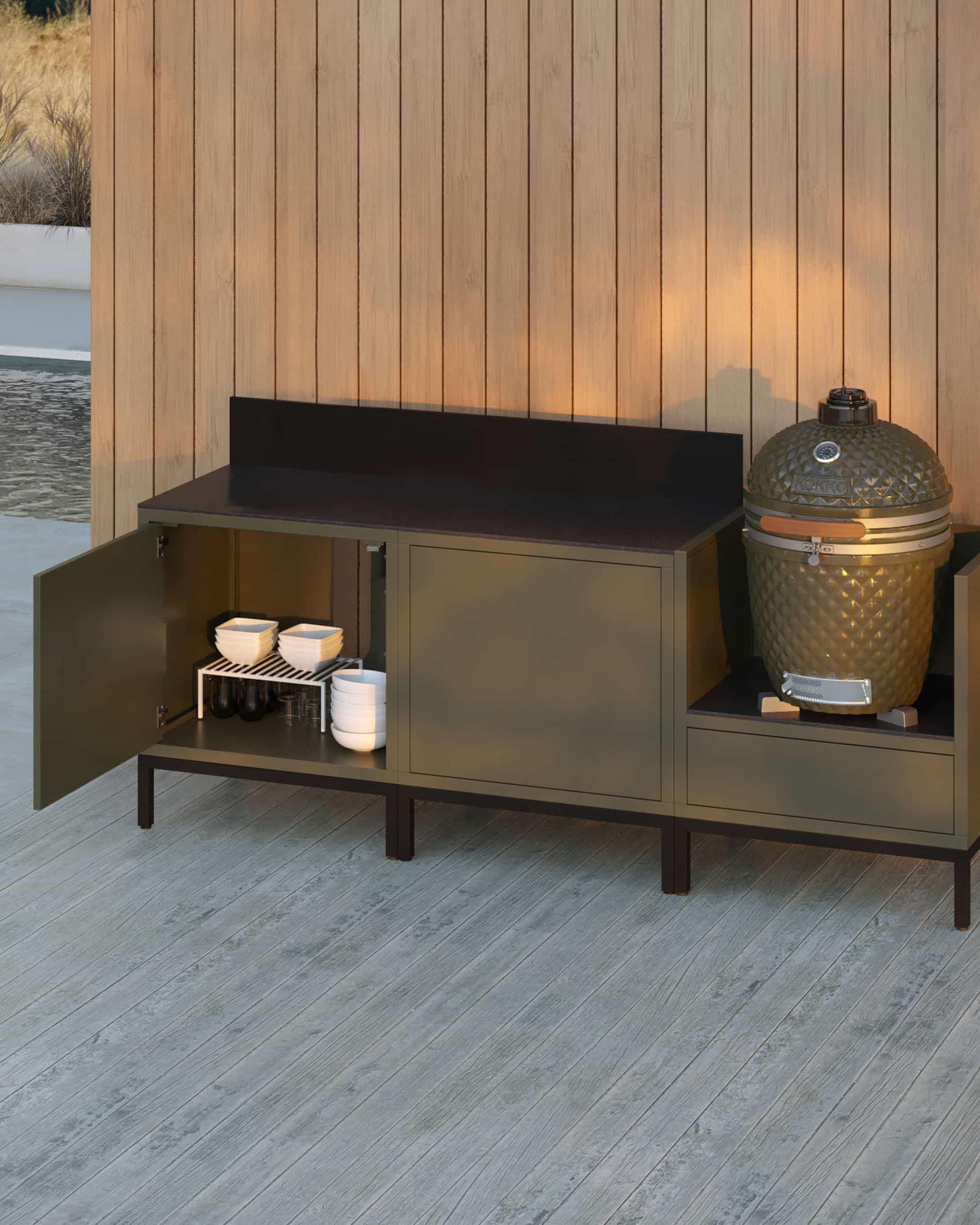 Buitenkeuken op palen met 3 modules voor kamado, meubel in olijfgroene kleur met donker werkblad en bijpassende achterwand, opbergdeur open met zichtbaar servies, kamado geplaatst in de rechter zijmodule, constructie op zwarte poten.