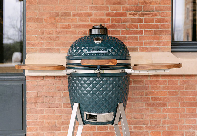 Kamado Kokko oceaanblauw op onderstel met houten zijtafels, van voren getoond buiten tegen een bakstenen muur.