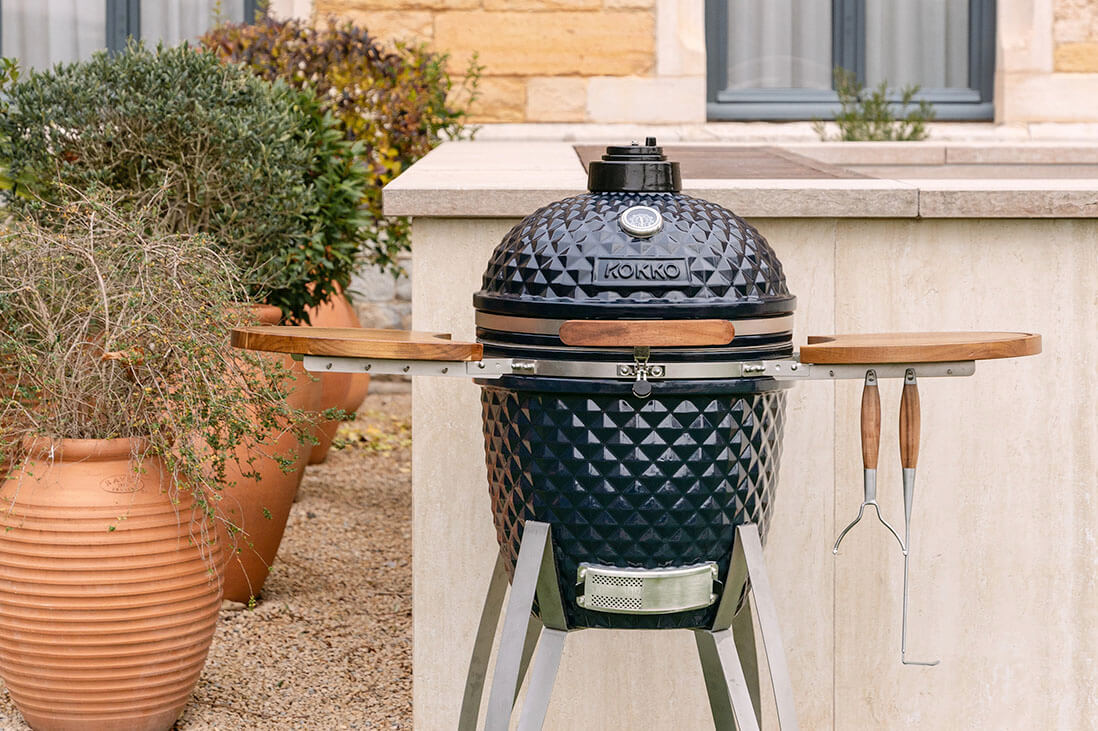 Kamado Kokko buiten opgesteld, met zwarte, getextureerde keramiek, houten zijtafels, hangende accessoires en metalen onderstel, geïntegreerd in een buitenkeuken.