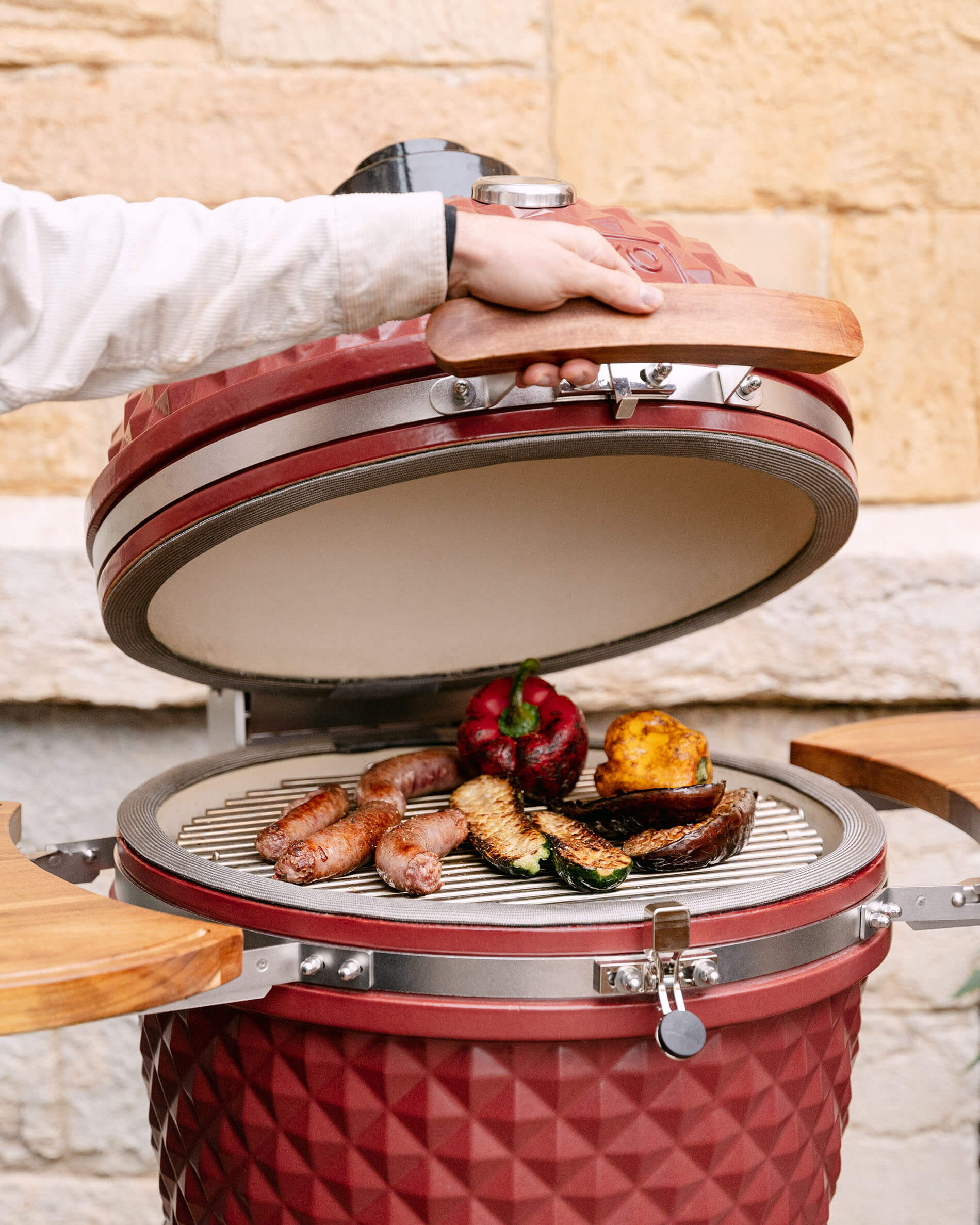 Open rode oxide keramische Kamado Kokko 22 inch, met worstjes en groenten die op het rooster worden bereid.